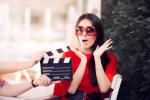 woman in a red shirt and red sunglasses looks surprised while another person uses a movie clapboard in front of them