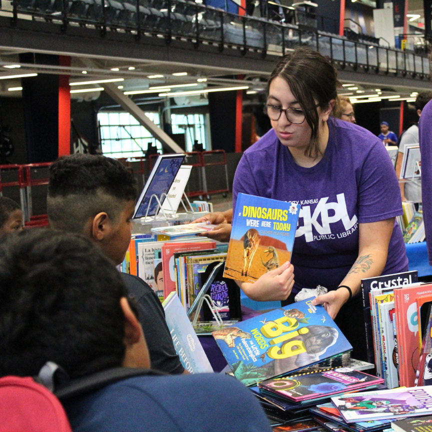 woman in a purple KCKPL shirt shows a dinosaur book to children from behind a table of books