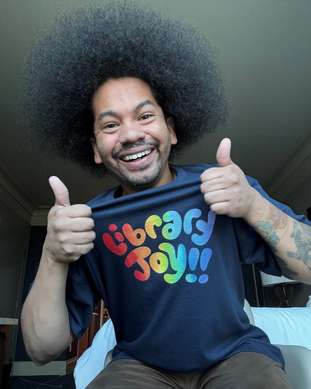 National Library Week honorary ambassador, Mychal Threets gives 2 thumbs up while wearing a dark t-shirt that reads Library Joy in rainbow lettering