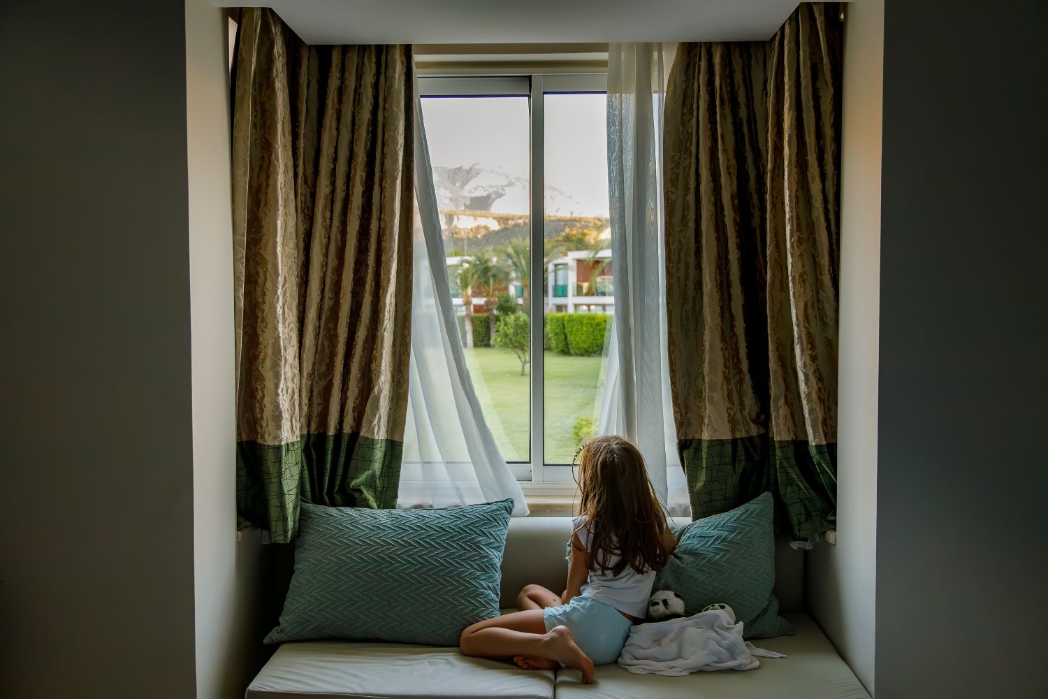 young girl looking outside sitting on a window bench with pillows