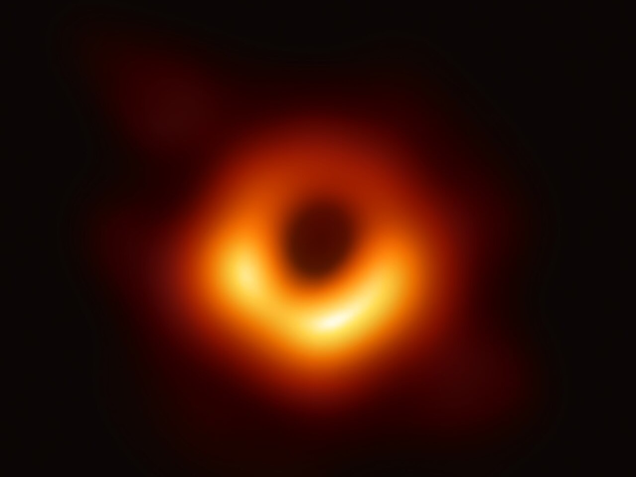 Image of the M87 supermassive black hole. It is an blurred orange and red swirl with a black spot in the center, surrounded by darkness.