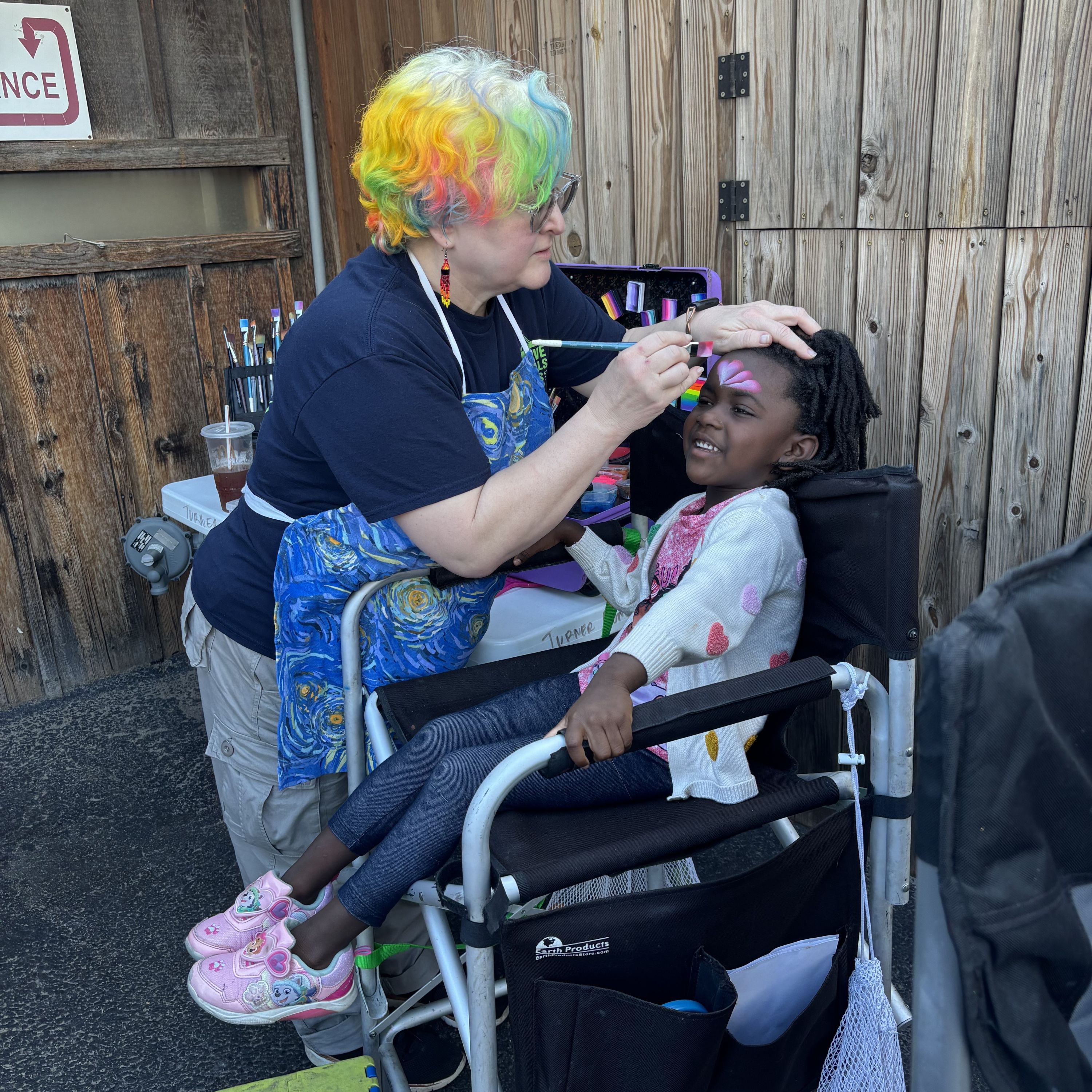 a librarian wearing a colorful clown wig does face paint on a young black girl 