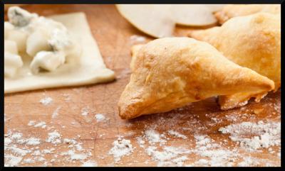 Triangular pastry sitting on a floured wood surface with a small square of raw pastry dough sitting to the side with chunks of cheese on it.