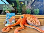an orange plush toy octopus holds onto the book "Remarkably Bright Creatures" in front of a fish tank