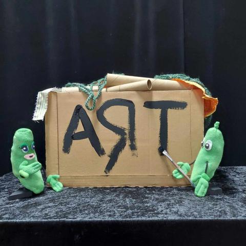 Two green pickle puppets ad a cardboard box labeled art. 
