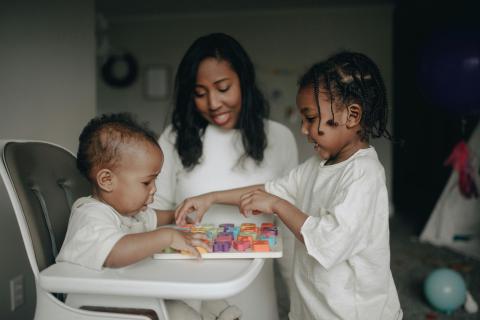A young women babysitting two children, a baby on the left in a high chair, and an older child with braids on the right.