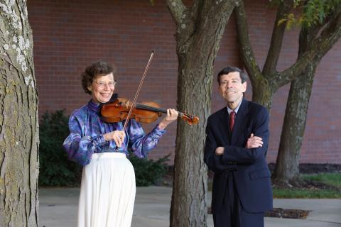 Picture of Susan and William Goldenberg. Susan is wearing a blue blouse and white skirt and playing a violin. William is wearing a dark blue suit and standing with arms crossed.