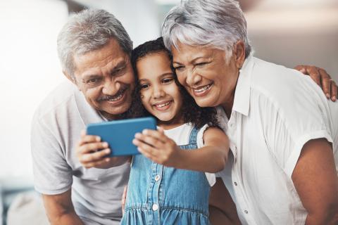 Small child in denim overalls holding a phone, taking a picture with a granddad and grandma