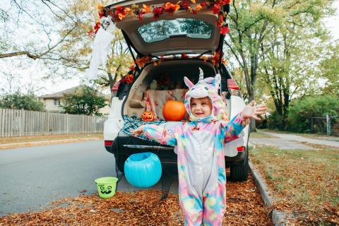 car trunk that is decorated for halloween with a child in a fuzzy unicorn costume standing in front of the car with a turquoise pumpkin trick or treat bag