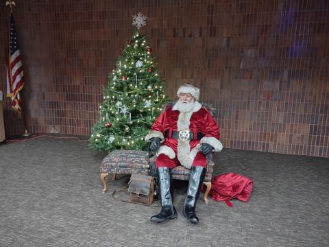 Santa with red coat and hat sitting in a wing chair next to a Christmas Tree