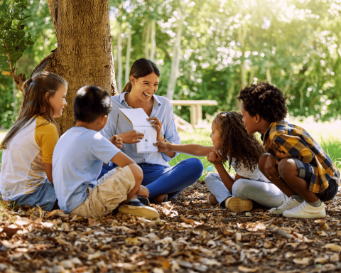smiling woman sitting under a tree showing 4 children a book