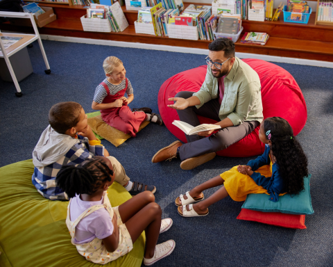 man sitting on a red cushion on the floor reads to children