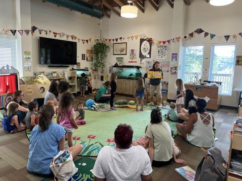 Librarian standing reading a book to a room filled with children and their parents around a large green carpet with lots of plants and couple of turtles behind the librarian