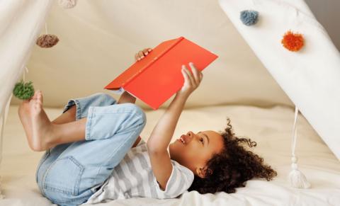 A child lies inside a tent reading a red book, with legs up in the air.