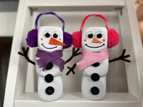 Two snowmen made from three foam dice stacked ontop of each toher decorated with eyes, scarf, ear muffs, 3 black buttons down center of belly, and brown pipe cleaners for stick hands.
