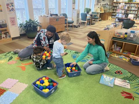 A librarian working with two children and an adult with colorful ball pit ball on the green carpet, with two books next to her that she will read.