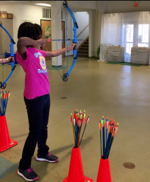 A child pulling back a bow with an arrow, next to some bright orange cones and arrows