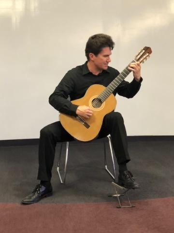 A man in all black sits on a chair on stage while playing acoustic guitar against a white wall background.