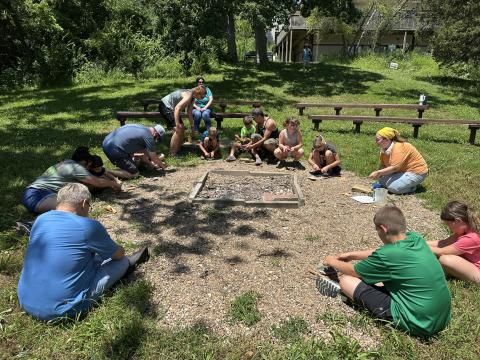 Children sitting around a fire pit in a circle with fire tools