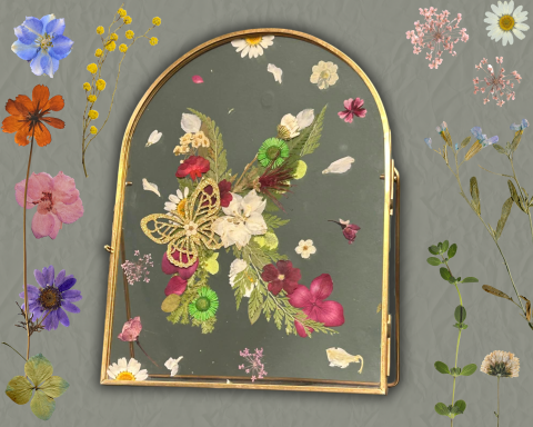 Gold framed transparent glass in the shape of an arch, adorned with pressed flowers and leaves that are arranged into the letter K. The arch frame is surrounded by pressed flowers and leaves of different colors on a green background.