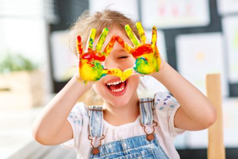 young child holds her paint covered hands in front of her face while smiling