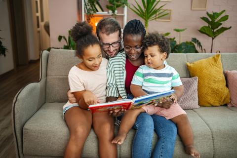 a family with an adult male, adult female, and 2 children reading a story on a couch. 