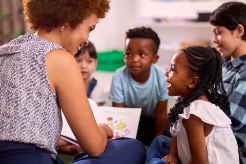 Female Teacher Reads To Multi-Cultural Elementary School Pupils Sitting On Floor In Classroom