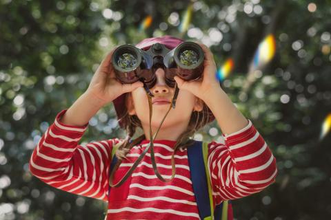 A child wearing a red and white striped, looking through a pair of binoculars, outside under the tree canopy.