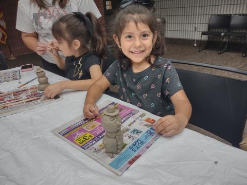 Small child with dark hair smiling and working on a clay piece at a table.