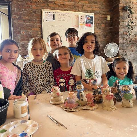 Seven children standing behind created clay pieces - sitting on a table.