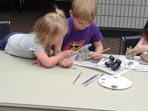 Two children with blond hair leaning on the table, sculpting a clay piece.