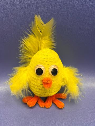 Egg-shaped chick with yellow feathers for wings, yellow pipe cleaner body, two eyes, an orange beak, and two orange pipe cleaner feet.