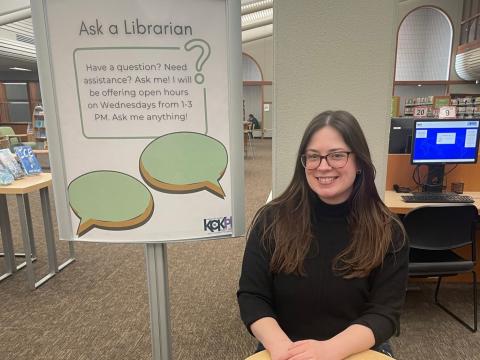 Katy, a librarian at West Wyandotte Library sits at a table in front of an ask a librarian sign