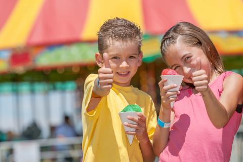 A young boy and girl are eating snow cones and giving a thumbs up at a carnival. 