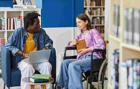 Two young adults, one in a wheelchair, conversing in a library setting.