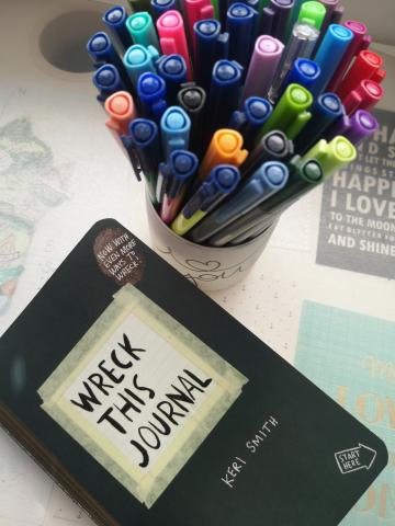 A black book titled Wreck This Journal in the bottom left corner with colorful markers in the top right corner