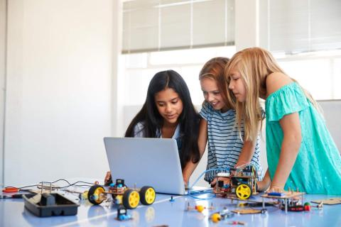 3 girls are looking at a laptop, with robotic pieces around them as they work together to code and build a robotic project. 
