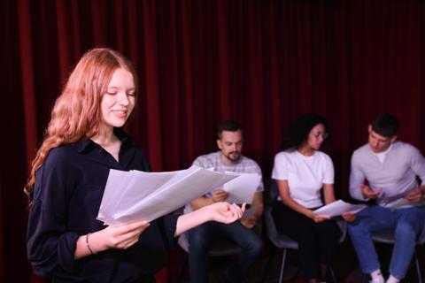 teen girl on stage at a theater reading from a script with other teens holding scripts seated behind her. 
