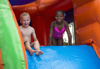 Little girl in pink swimsuit on hands and knees, smiling. Little boy sitting smiling. Both at top of an inflatable slide.