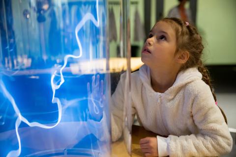 A girl is looking up as she is fascinated by an electrical experiment that shows of blue bolts of electricity. 