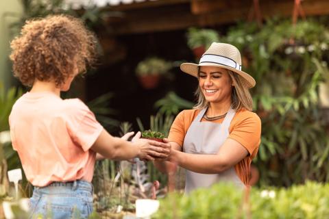 two women in a garden area with one handing another a small plant