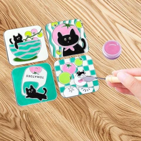 Four enamel pins on a wooden surface. A hand is working on adding the enamel paint onto the sections of the lowest right pin. Each pin has different cat and tomatoes designs. 