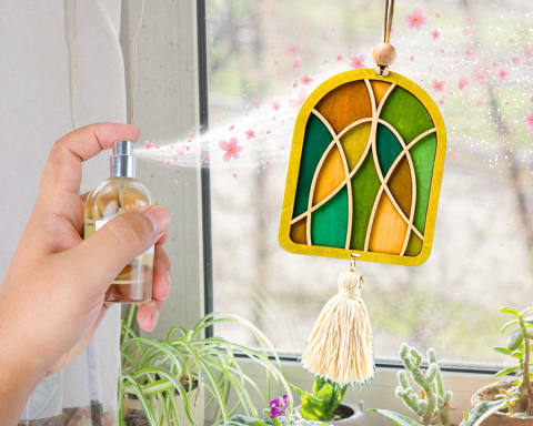 A wooden ornament is painted different colors as it hangs from a window as a tassel swings form it. A person is spraying a scent on the ornament to give it a floral smell.