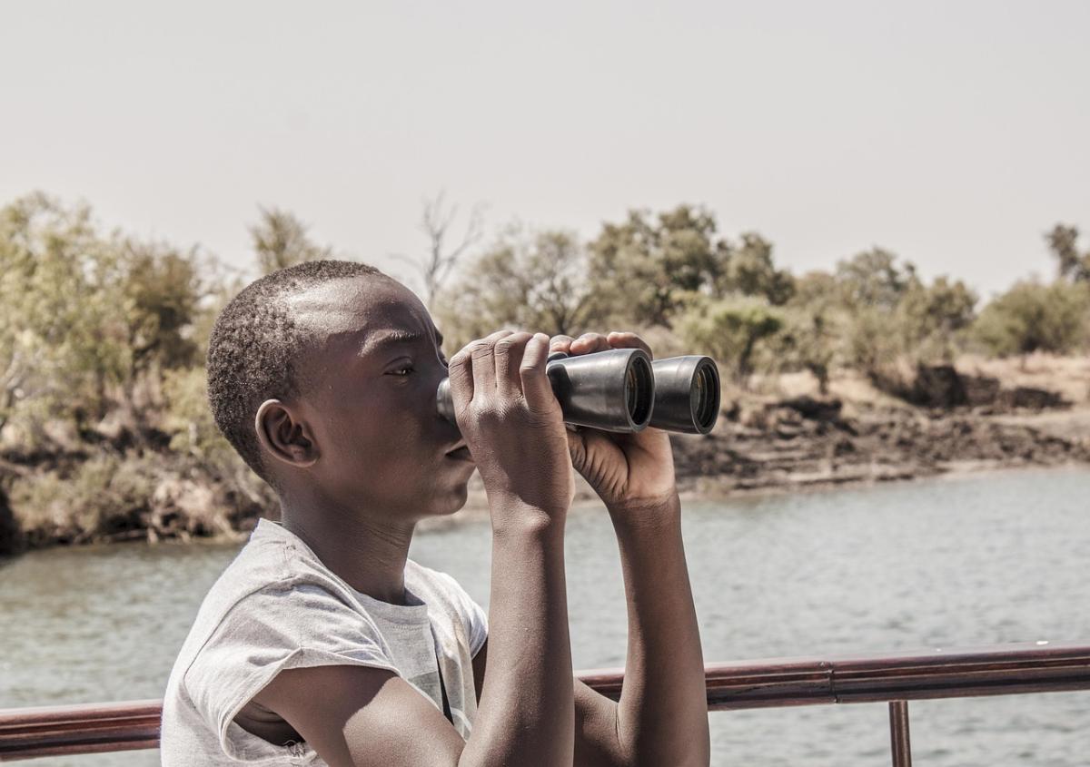 Child with darker complexion look in binoculars, near a lake, outside. 