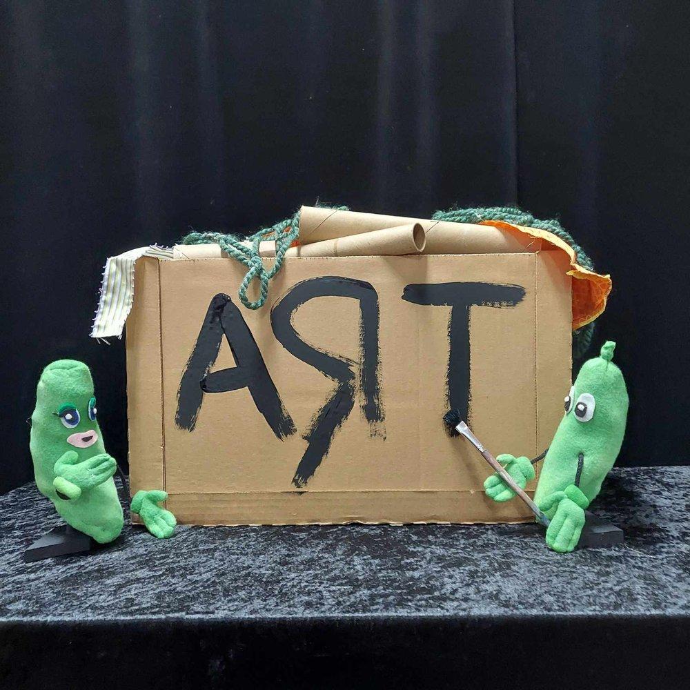 Two green pickle puppets ad a cardboard box labeled art. 