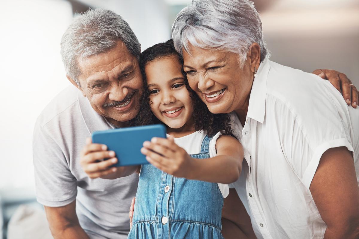 Small child in denim overalls holding a phone, taking a picture with a granddad and grandma