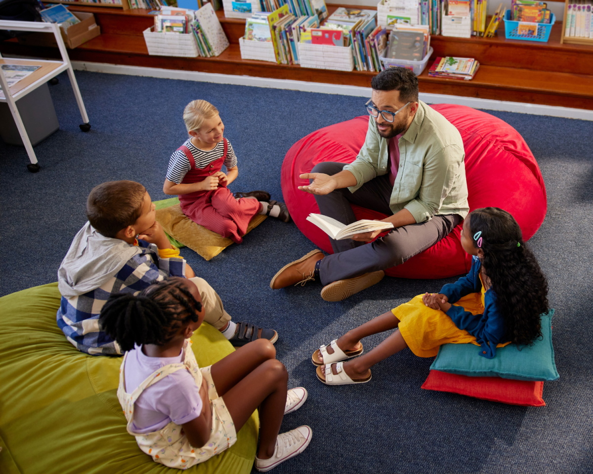 man sitting on a red cushion on the floor reads to children