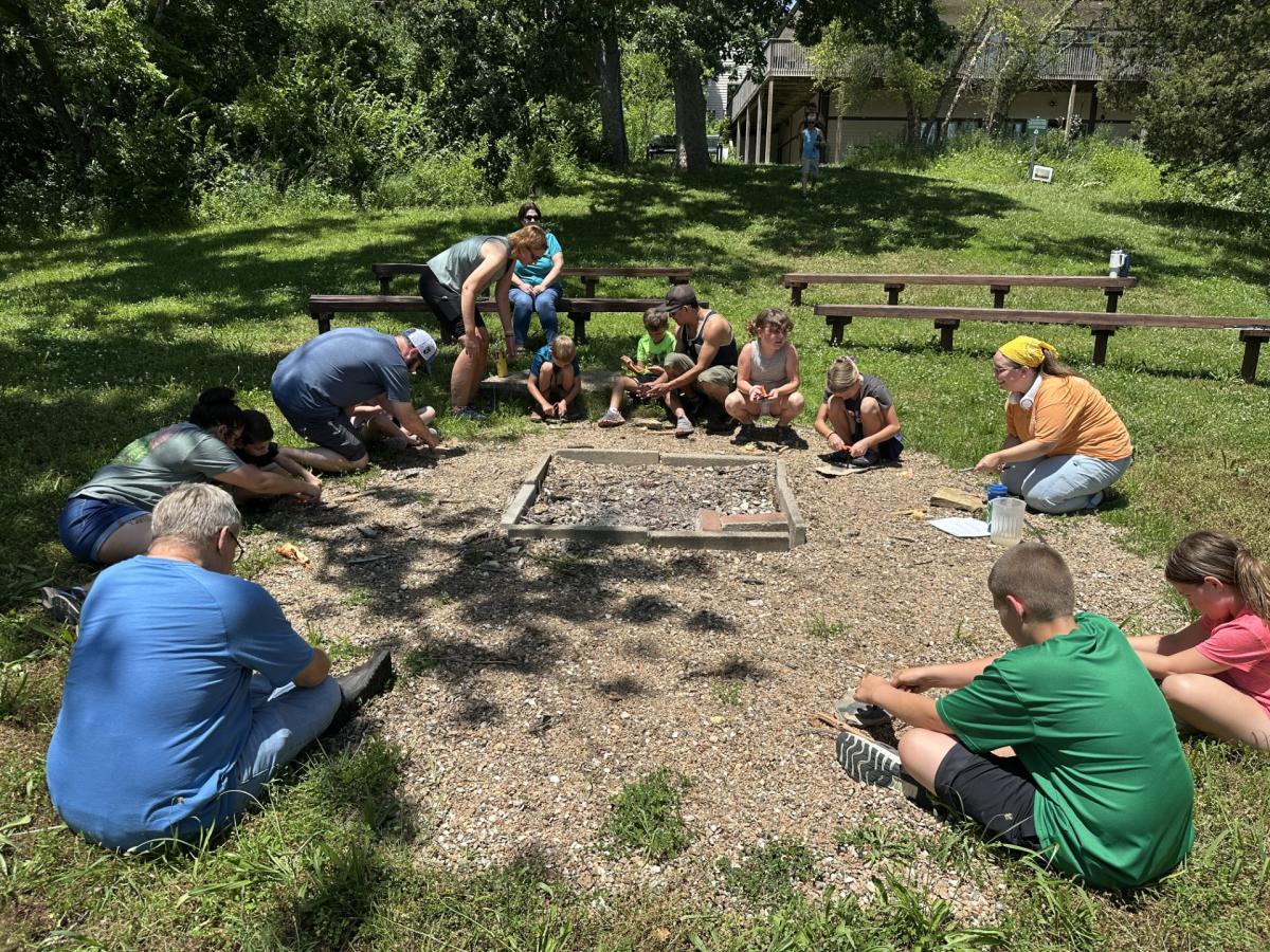 Children sitting around a fire pit in a circle with fire tools