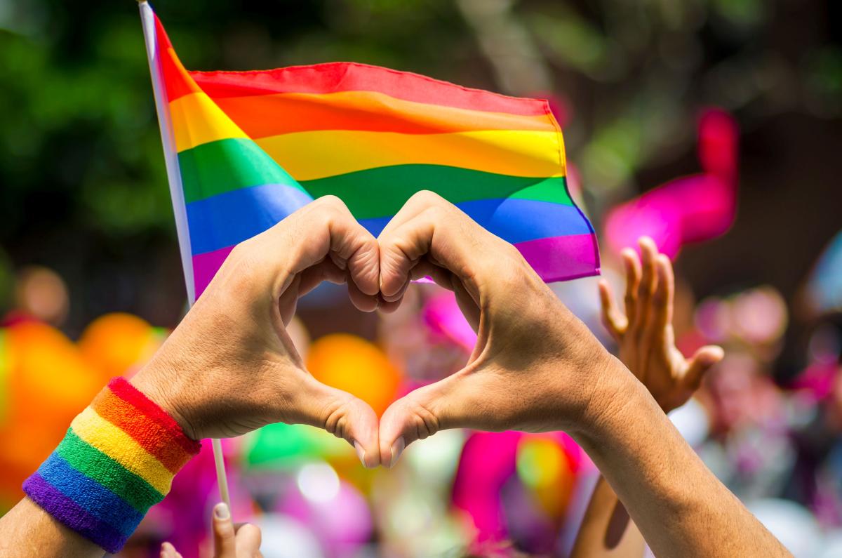 person holds up hands in heart symbols in front of a PRIDE flag