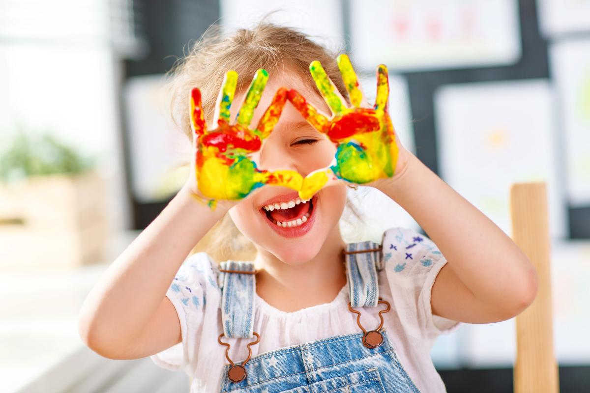 young child holds her paint covered hands in front of her face while smiling
